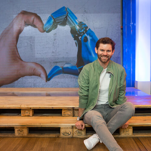 Der Politiker Florian Braun sitzt auf einer Palette vor einem Wandbild, auf dem eine menschliche Hand und eine Roboter-Hand gemeinsam ein Herz formen